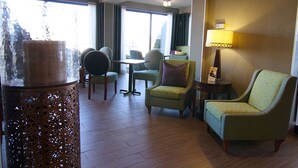 A hotel room with a wooden floor, a round table, and two chairs. There is a lamp on a side table and a large window with curtains. - Hampton Inn Winchester-University/Mall Area (Winchester)