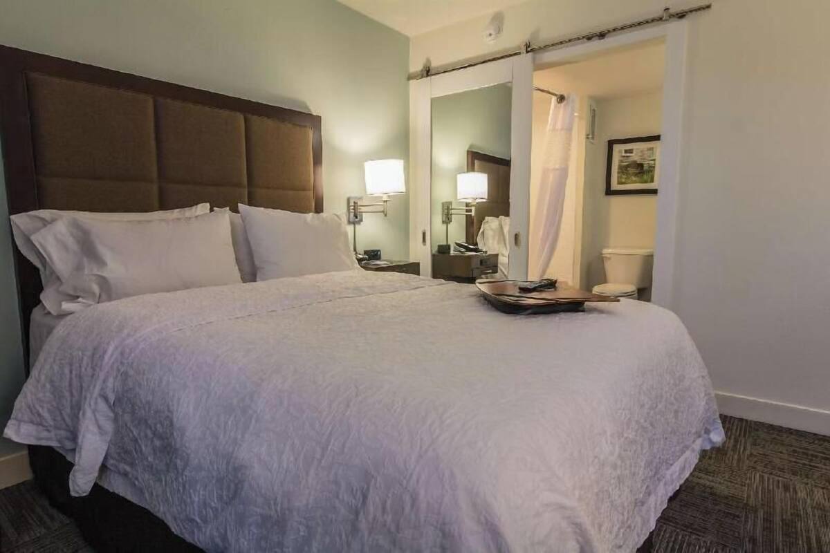 A hotel room with a large bed, a nightstand with a lamp, a bathroom door, and a framed picture on the wall.