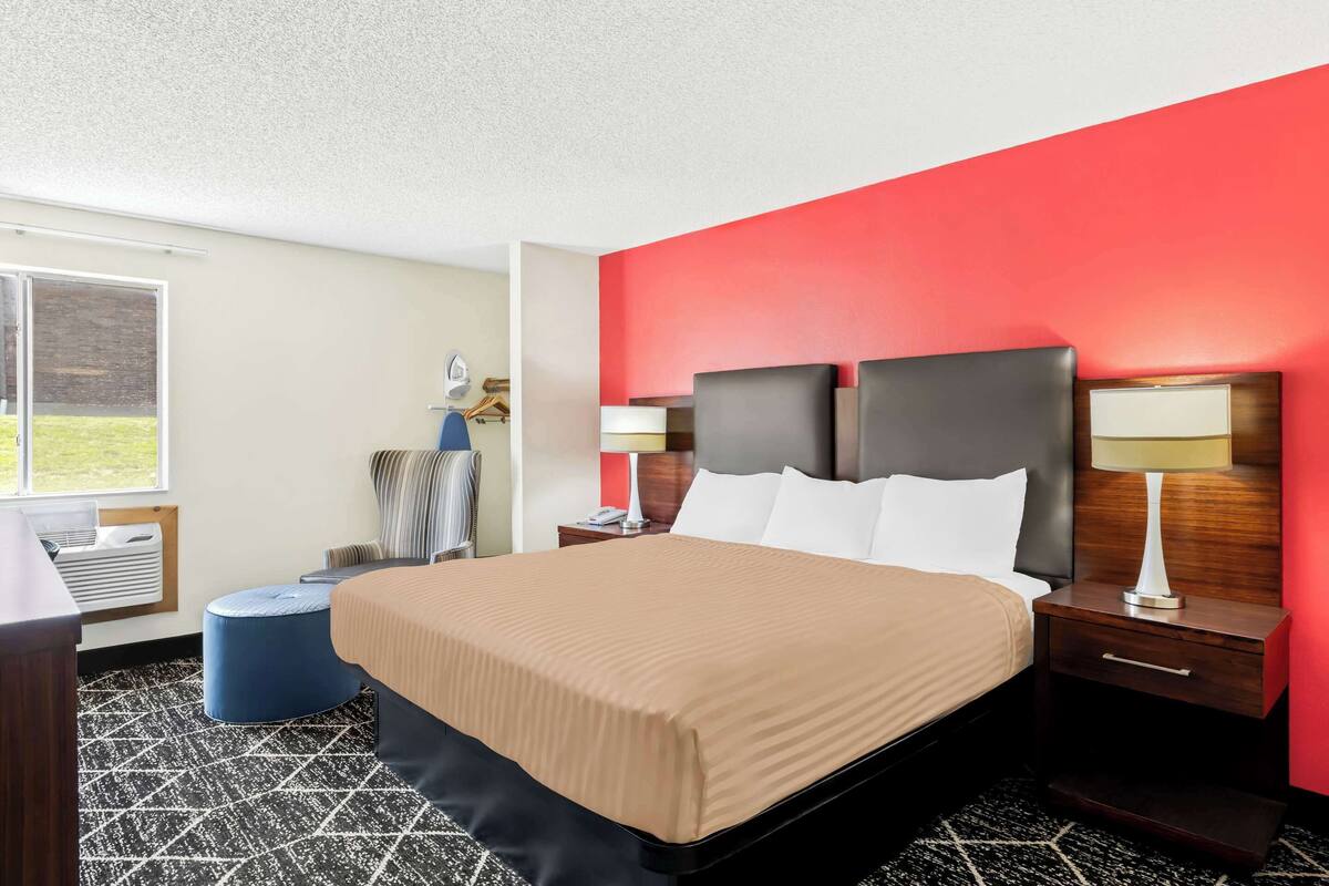 A hotel room with a large bed, a nightstand with a lamp, a blue ottoman, and a window with a view of the outdoors.