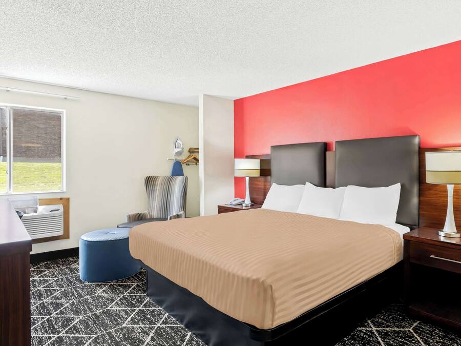 A hotel room with a large bed, a nightstand with a lamp, a blue ottoman, and a window with a view of the outdoors.