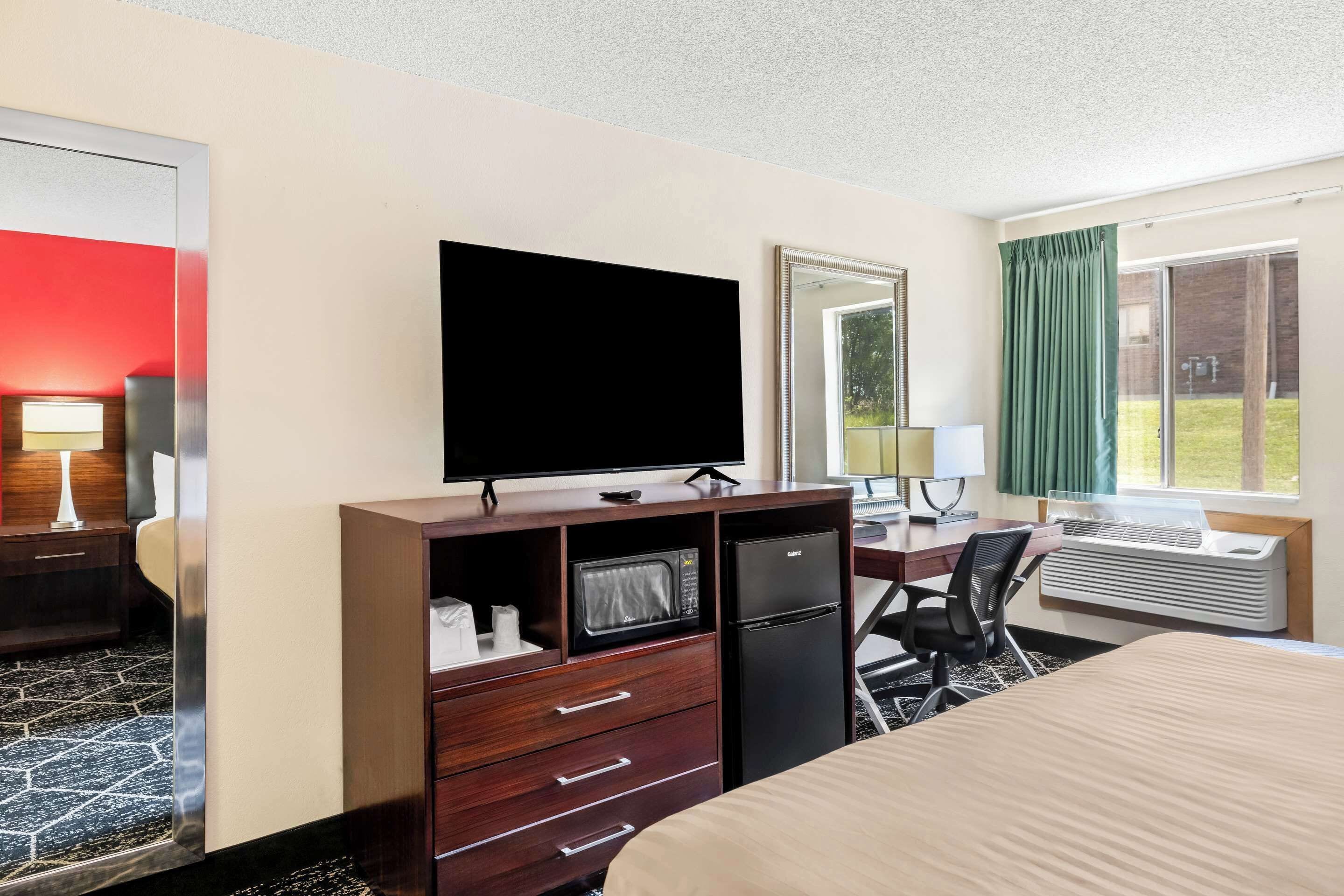 Habitación de hotel con cama, escritorio con silla, televisor, microondas, refrigerador y una ventana grande con cortinas.