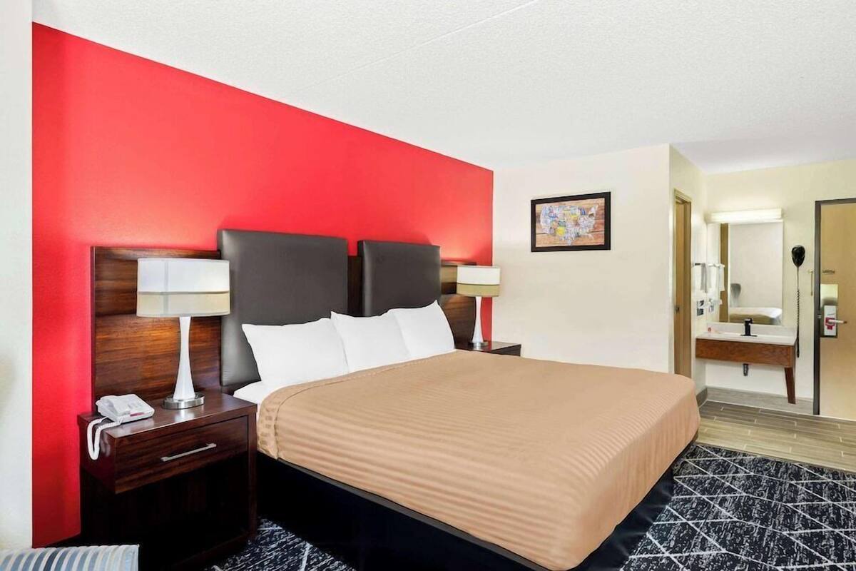 A hotel room with a bed, nightstands, a red accent wall, a painting, and a bathroom visible through an open door.
