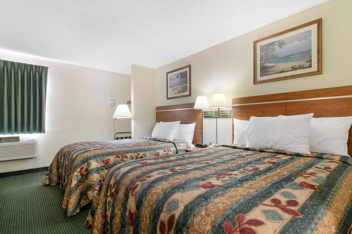 A hotel room with two beds, a window with curtains, a painting on the wall, and a lamp beside each bed.