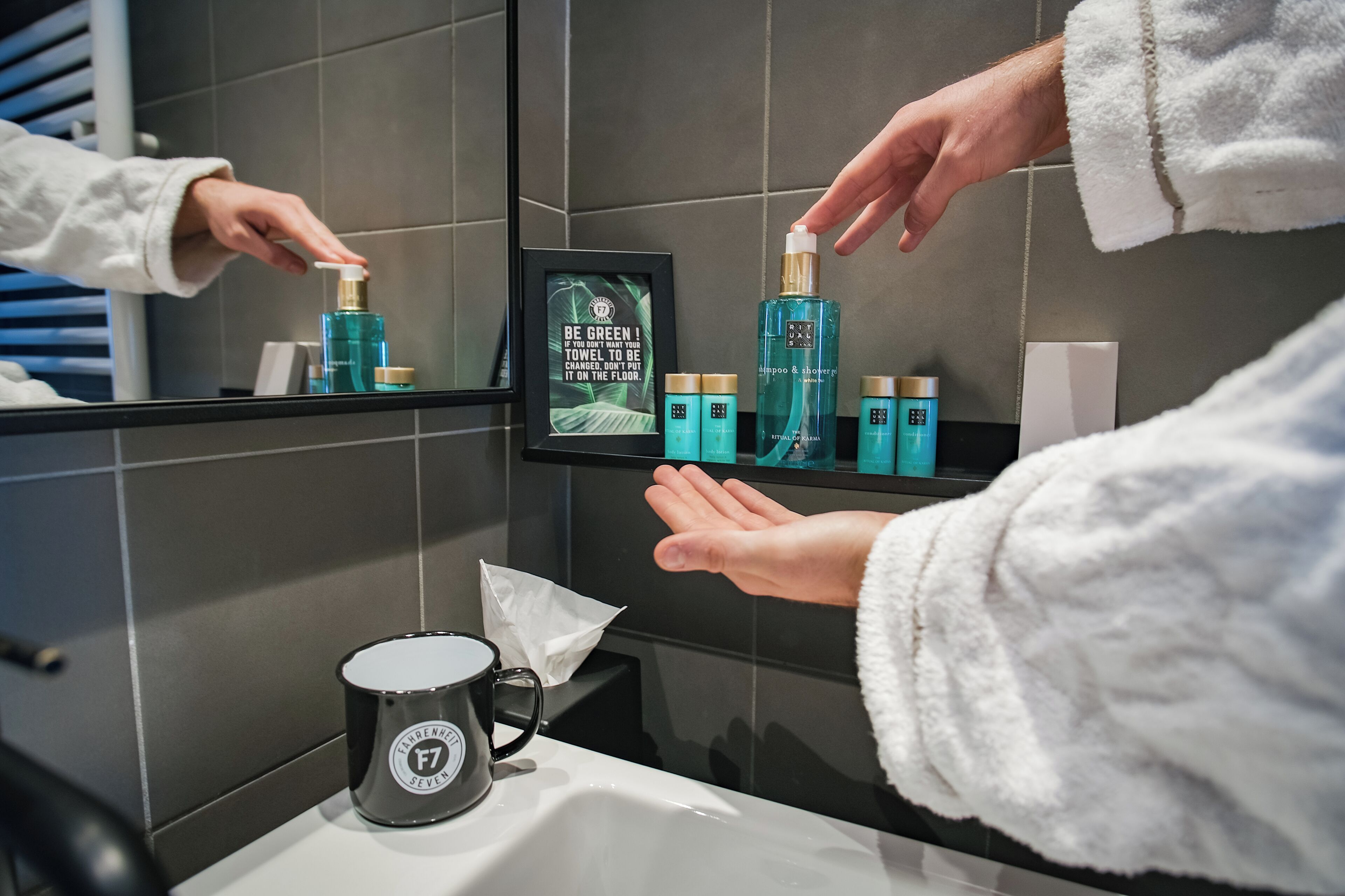 Une salle de bain avec un miroir, une étagère contenant des articles de toilette, un cadre avec une citation, et une tasse noire.