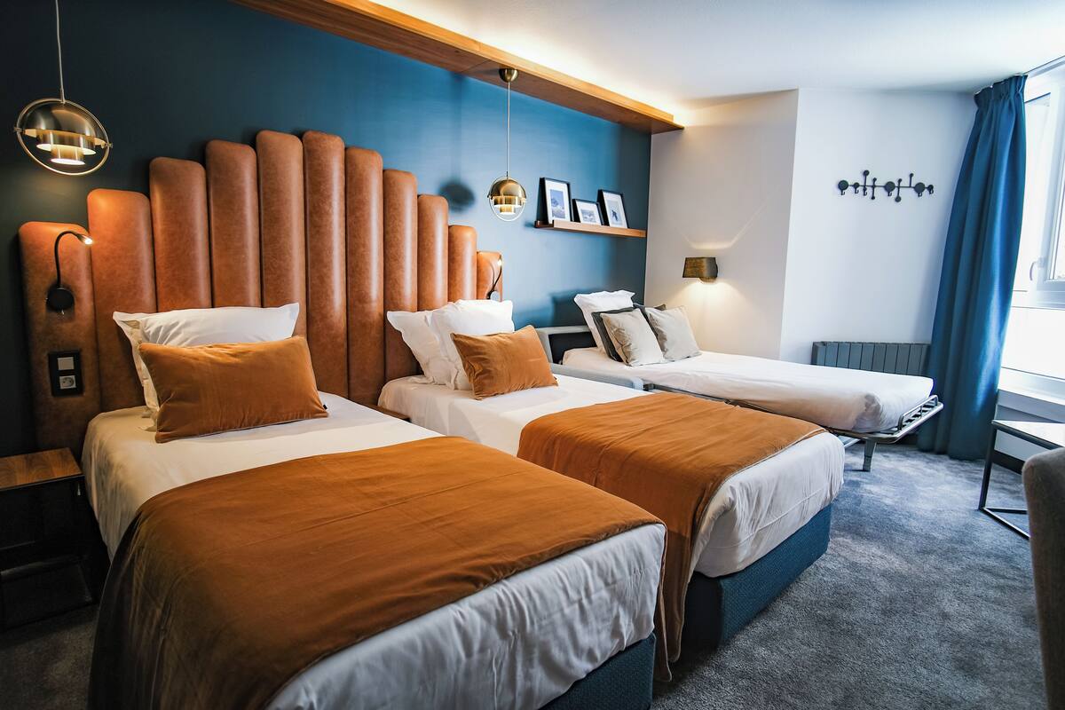 Ein Hotelzimmer mit zwei Betten, einem hölzernen Kopfteil, einem Regal mit Bildern und einem Fenster mit Vorhängen.