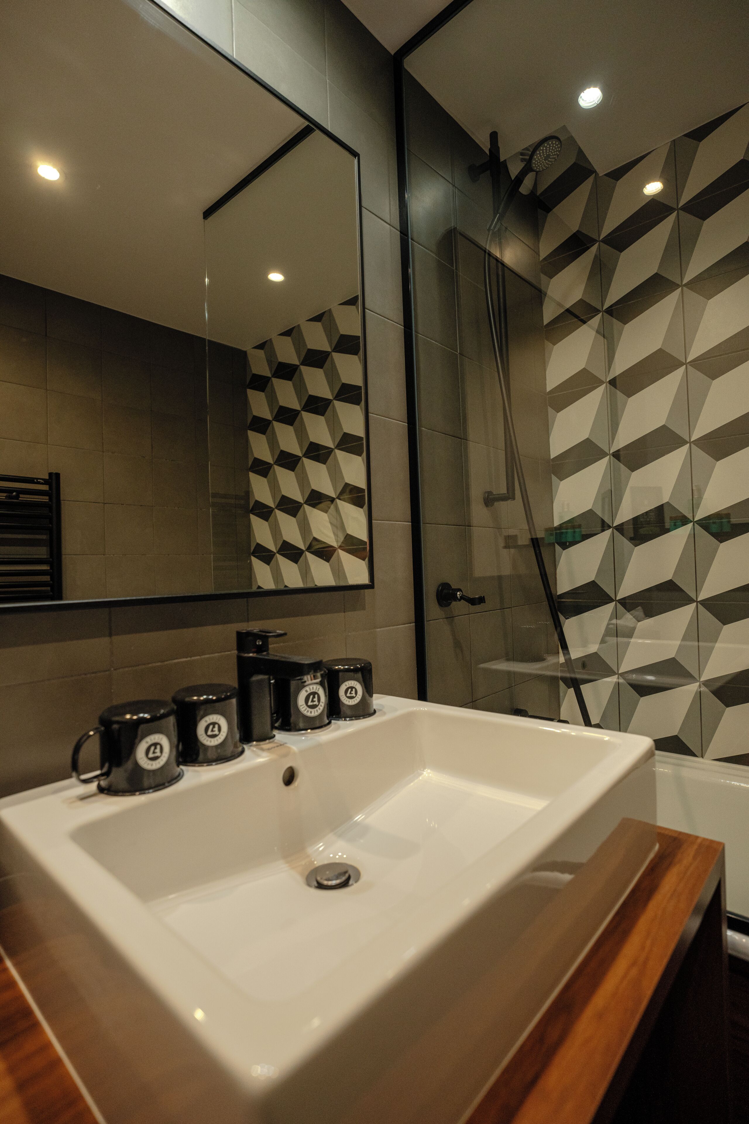 Ein Badezimmer mit großem Spiegel, weißem Waschbecken, Dusche und Fliesen mit geometrischem Muster.