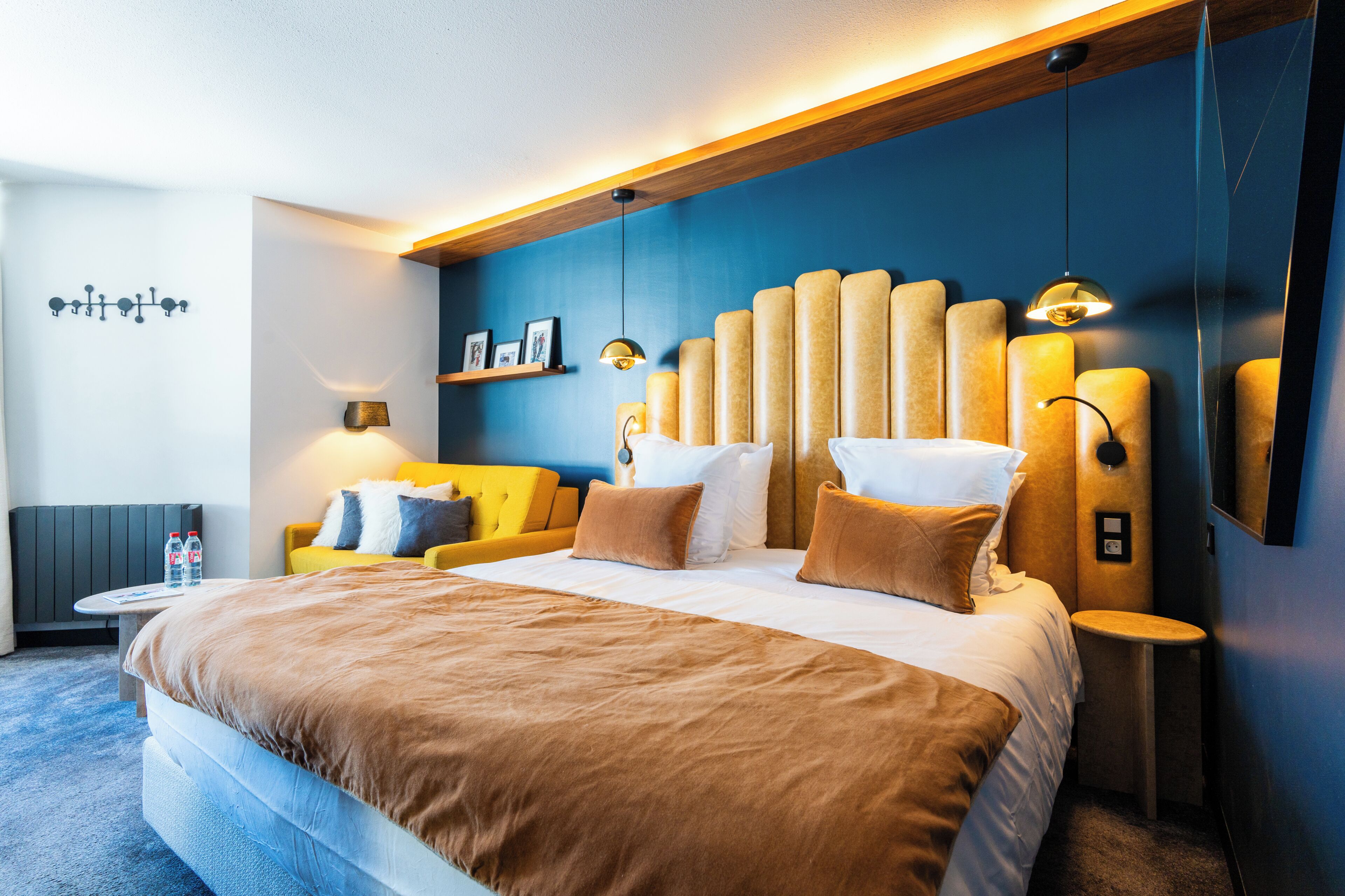 A modern hotel room with a large bed, a sofa, a small table, and a wall-mounted shelf with pictures.