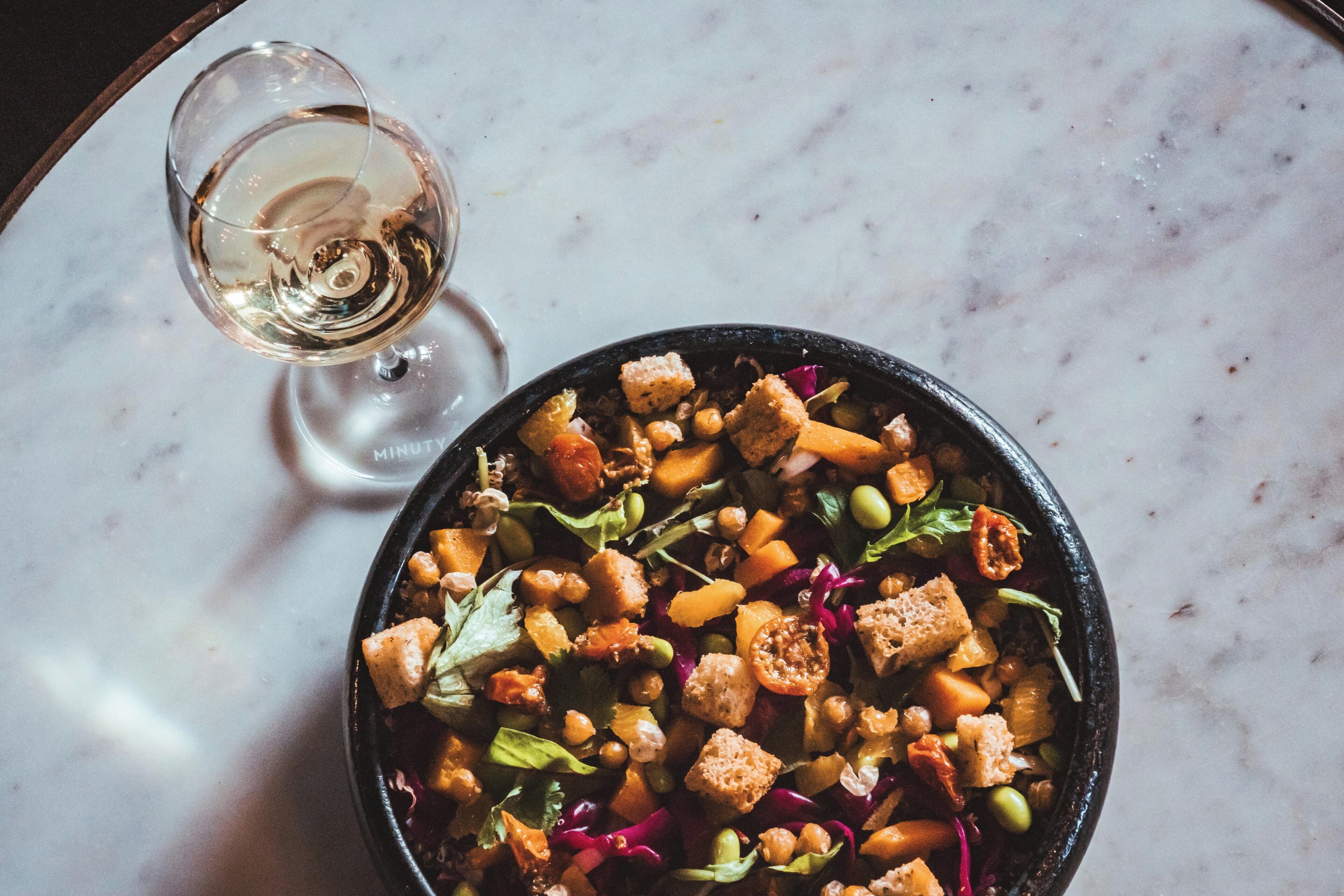 A bowl of salad with croutons and chickpeas on a marble surface.