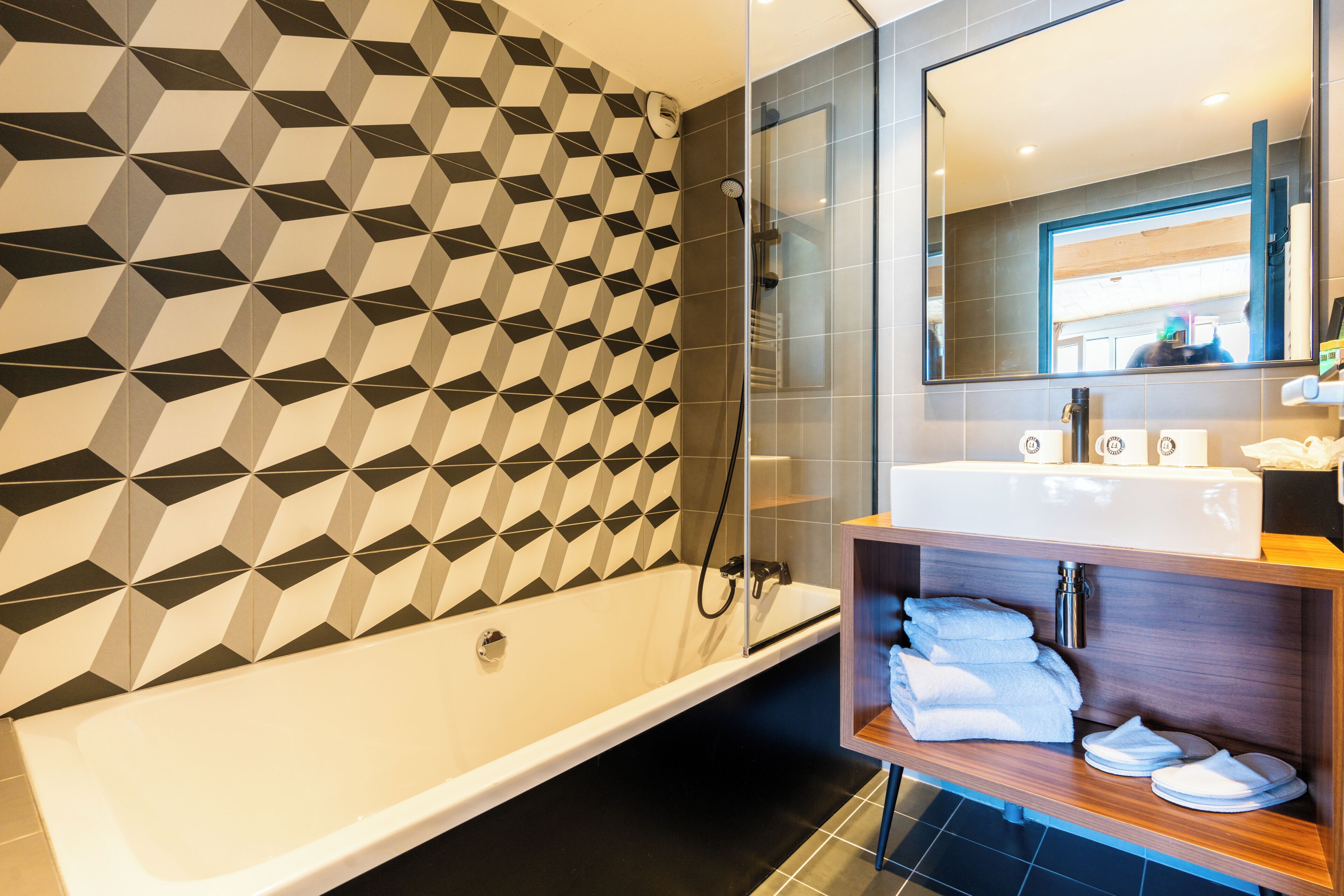 A modern bathroom with a geometric patterned wall, a freestanding bathtub, a wooden vanity with towels, and a large mirror above the sink.
