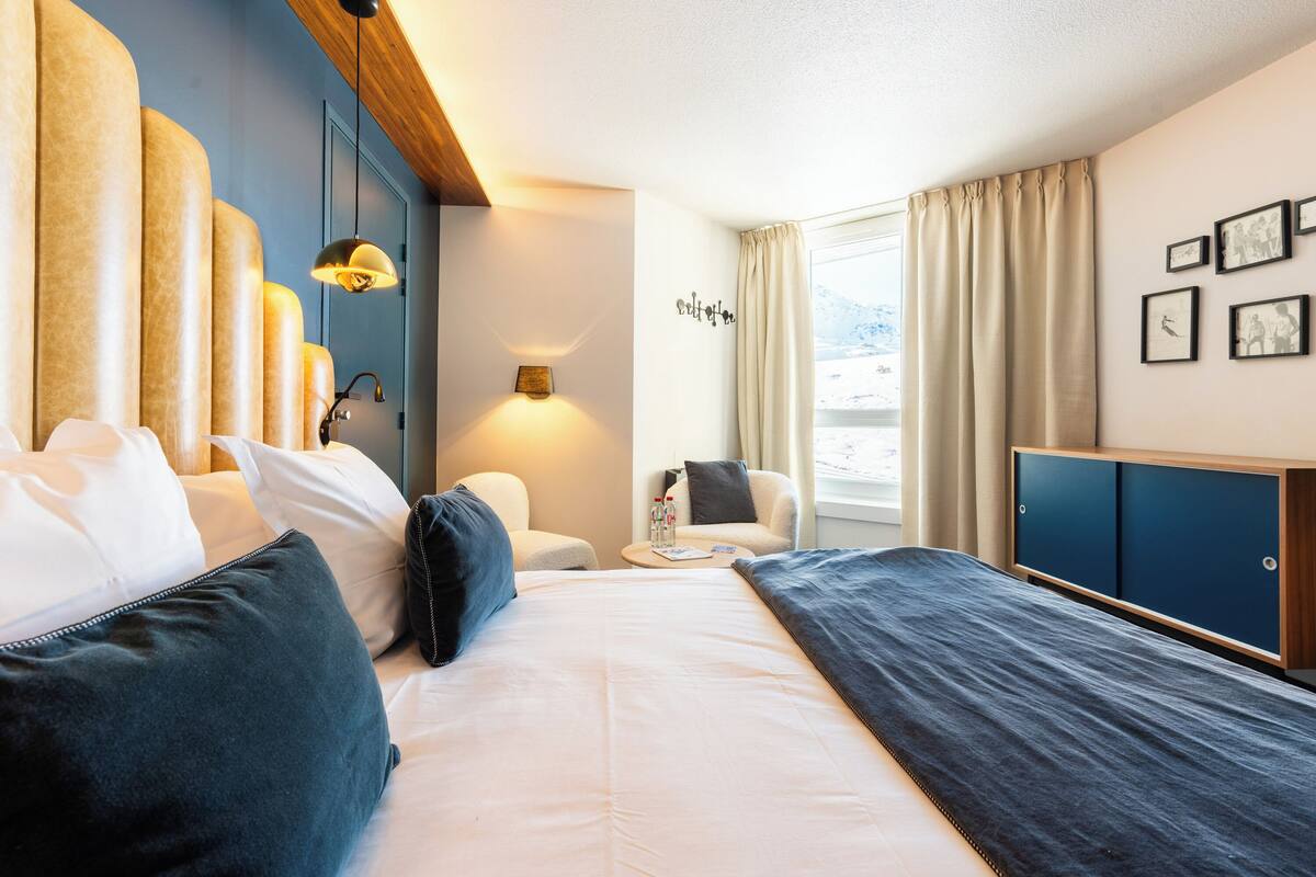 Ein Hotelzimmer mit Bett, Sessel, Nachttisch, Kleiderschrank und einem Fenster mit Vorhängen.