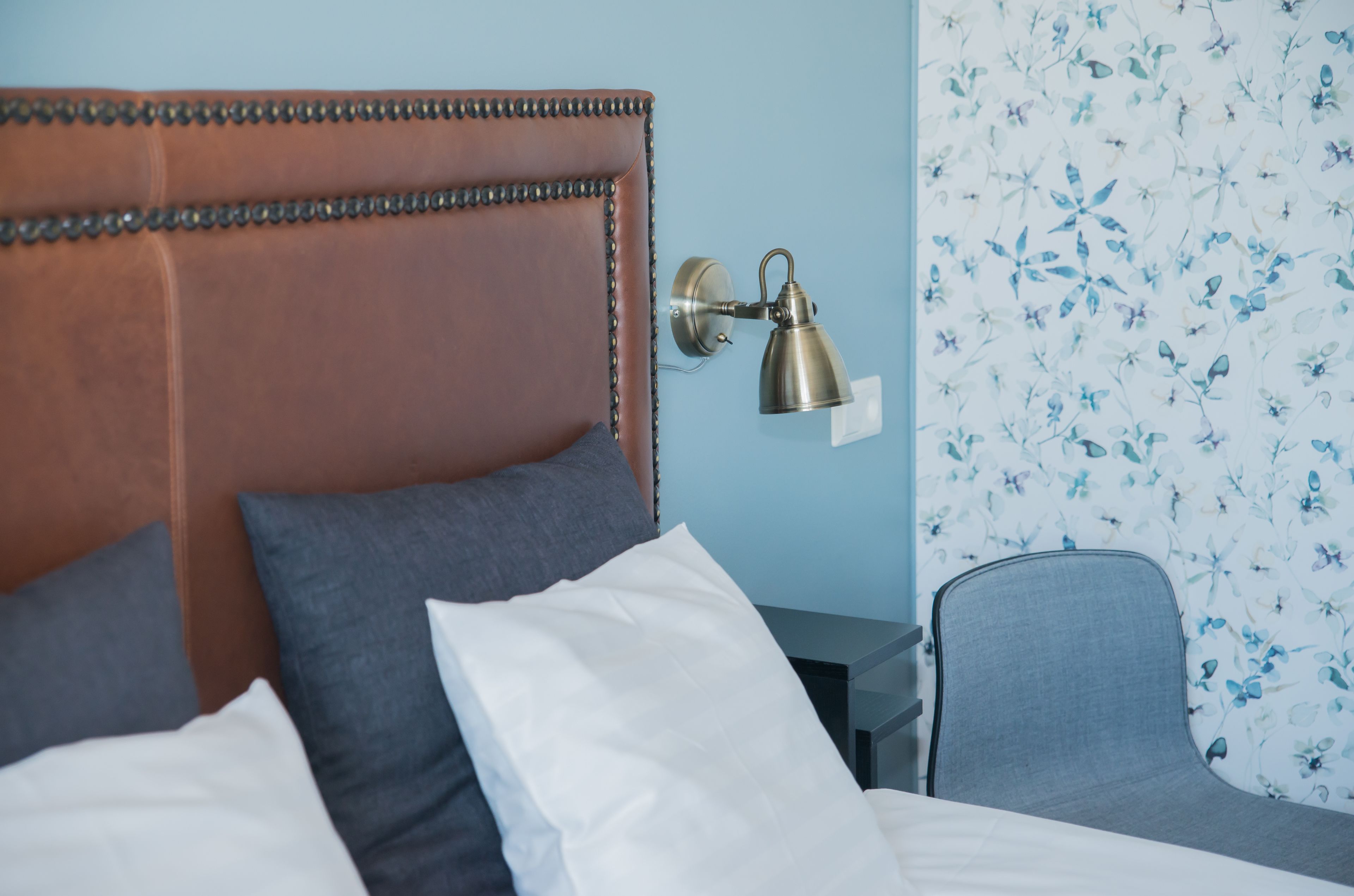 A neatly made bed with a brown headboard, a bedside lamp, and a chair with a floral wallpaper background.
