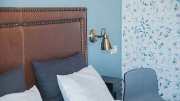 A neatly made bed with a brown headboard, a bedside lamp, and a chair with a floral wallpaper background.