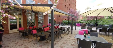 An outdoor patio area with tables and chairs, covered by umbrellas, and surrounded by brick buildings.
