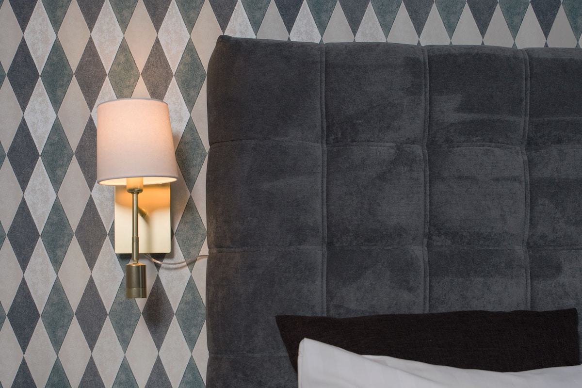 Un lit bien fait, avec un oreiller blanc et une tête de lit sombre. Une table de chevet avec une lampe est visible contre un mur à motifs.