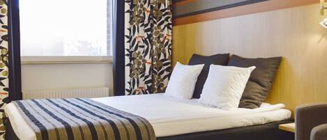 A hotel room with a bed, a window with blinds, a curtain with a leaf pattern, and a wall with horizontal stripes.