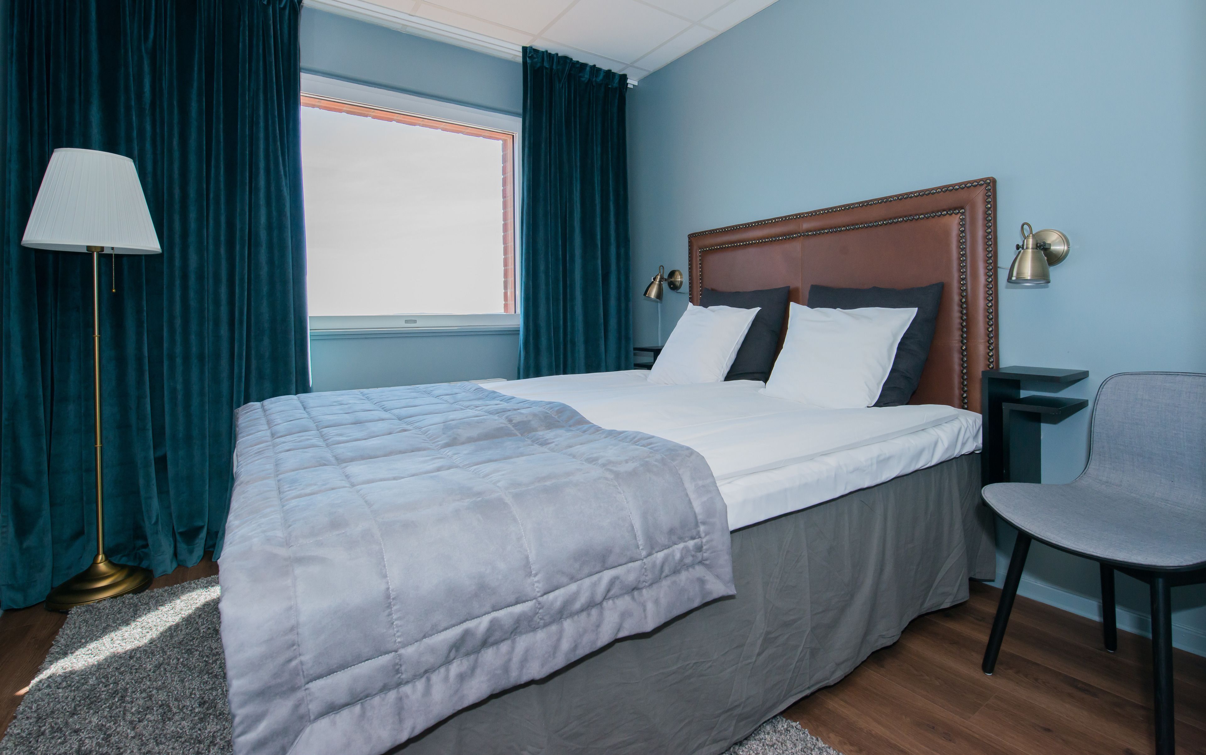 Una cama bien hecha con edredón gris, cabecera de madera y lámpara de noche.