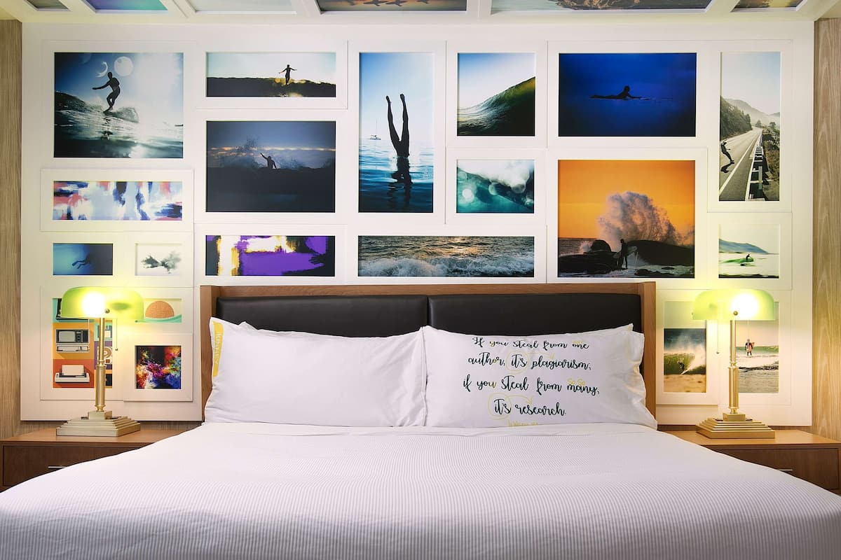 A hotel room with a large bed, two bedside tables with lamps, and a wall decorated with framed photographs of landscapes and seascapes.