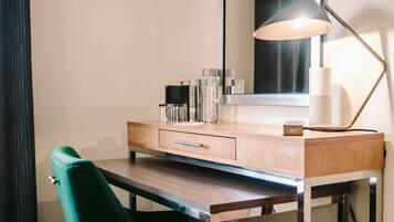 A hotel room with a wooden desk, a green chair, a large mirror, and a floor lamp.
