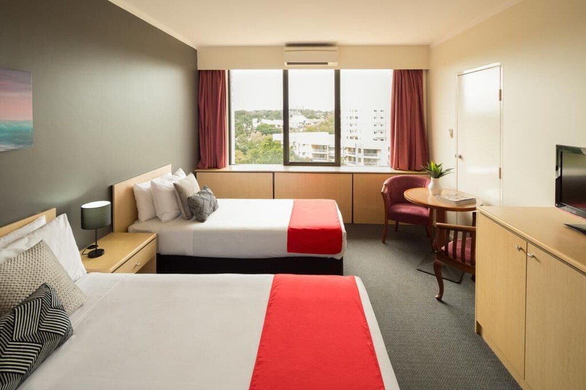 A hotel room with two beds, a desk, a chair, a TV, and a window with a city view.