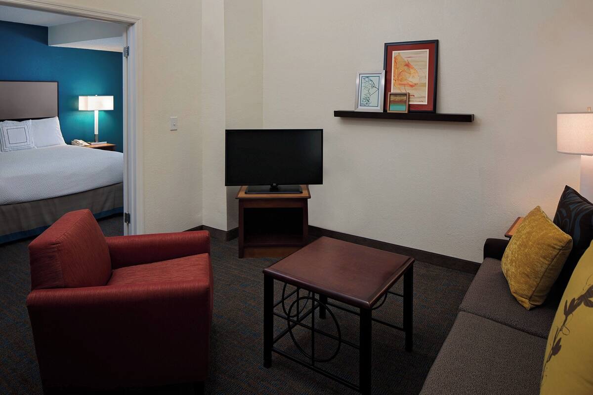 A hotel room with a bed, a red armchair, a small table, a TV on a stand, and a sofa with a yellow pillow.