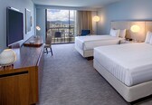 A hotel room with two beds, a desk with a flat-screen TV, a chair, a view of the cityscape, and a balcony with curtains.