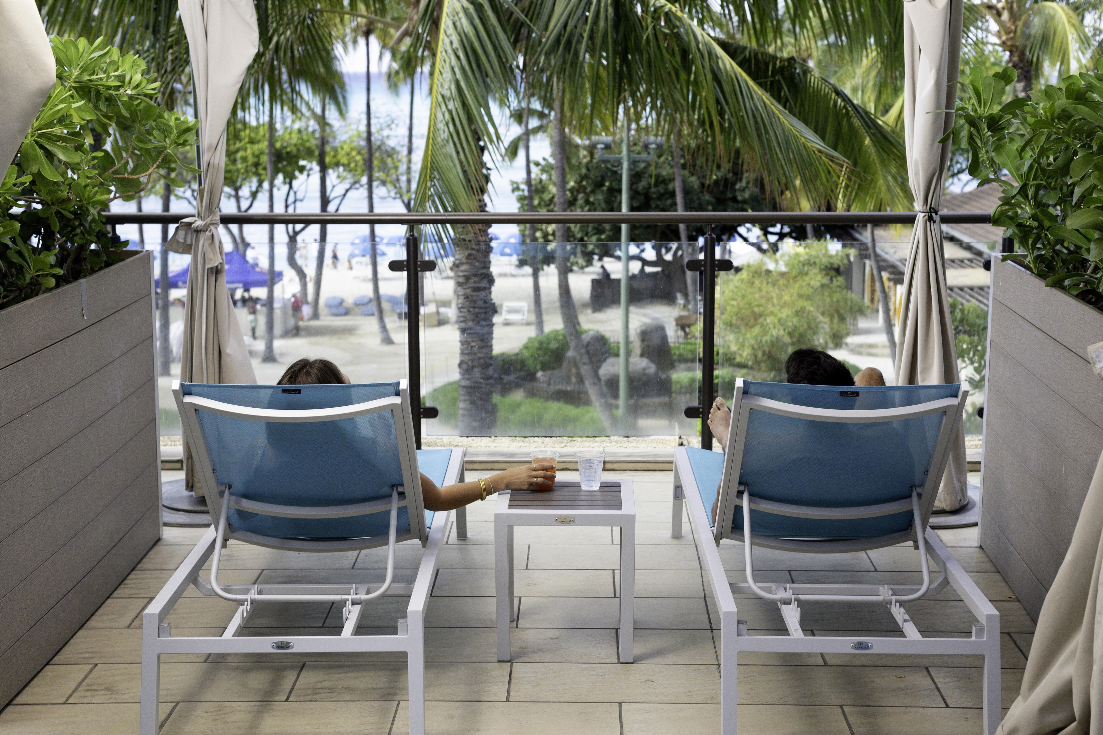 Two lounge chairs with a table, a view of palm trees and beach, and a railing.
