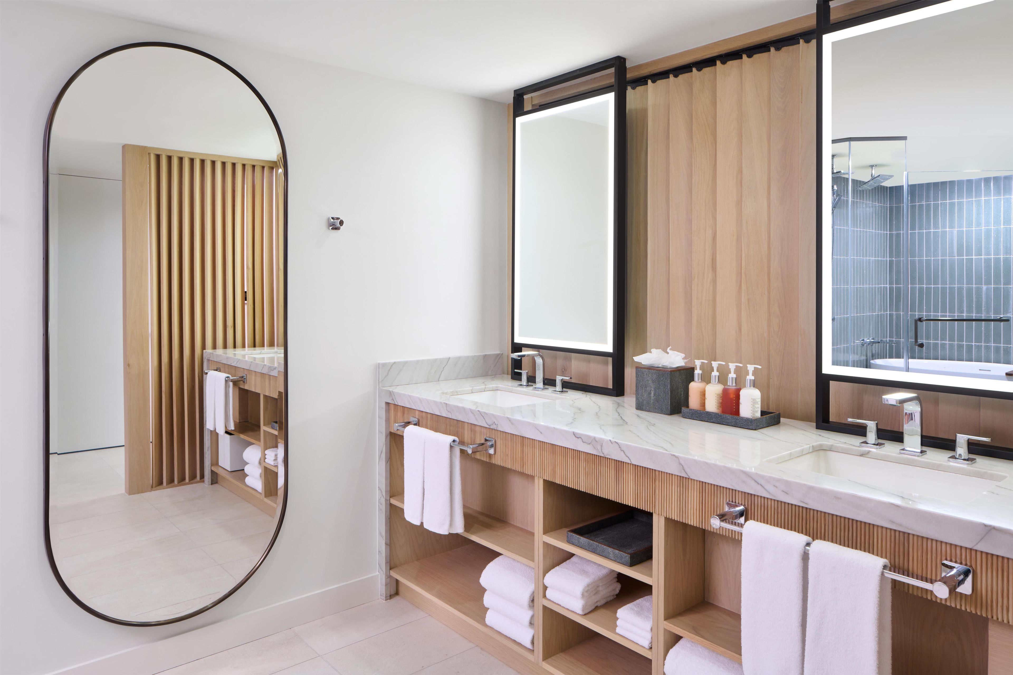 Un baño moderno con un espejo grande, dos lavamanos, estantes de madera y una zona de ducha con azulejos.