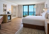 A hotel room with a large bed, a desk, a chair, a small table, and a view of the ocean.