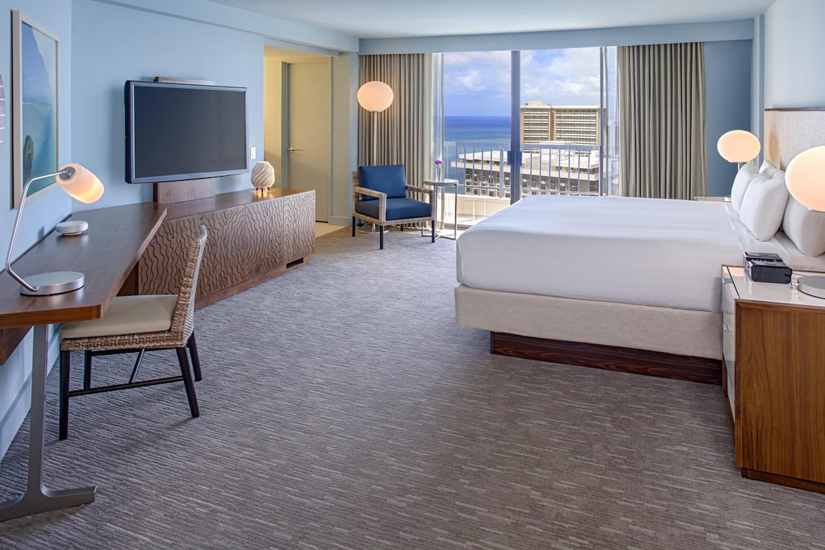 A hotel room with a bed, desk, chair, TV, and a view of the ocean.