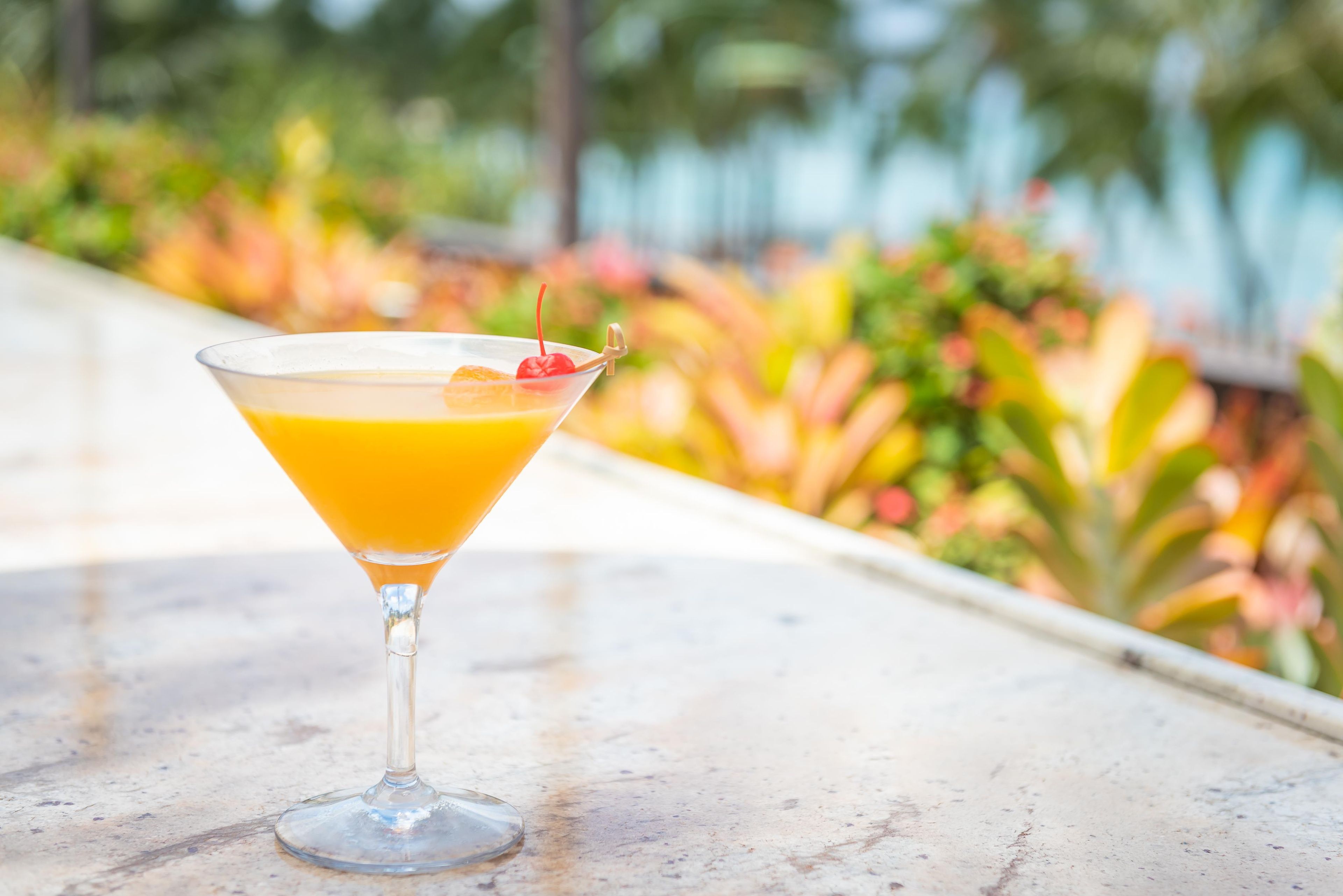 A martini glass with a yellow-orange drink on a marble surface, outdoors with a blurred garden background.