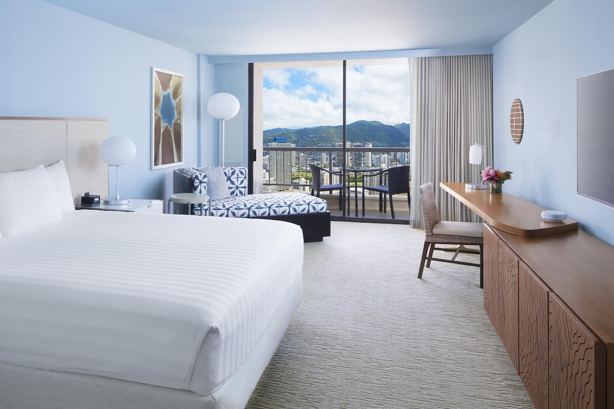 Ein Hotelzimmer mit einem großen Bett, einem Schreibtisch, einem Sessel, einem Balkon mit Blick auf die Stadt und einem großen Fenster.
