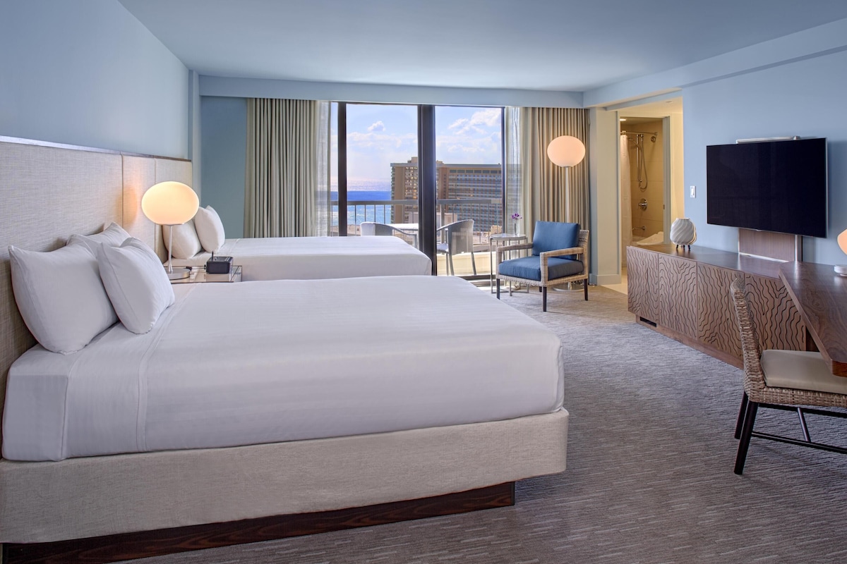 Ein Hotelzimmer mit zwei Betten, einem Schreibtisch, einem Sessel, einem Fernseher und Blick auf die Stadt.