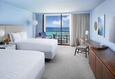 A hotel room with two beds, a desk, a chair, a TV, and a view of the ocean.