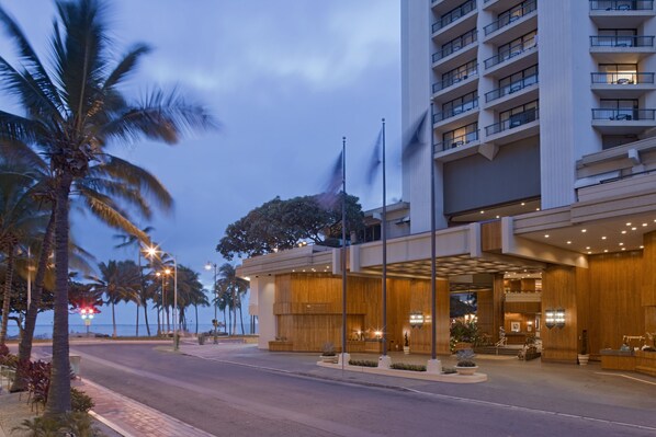 Un hôtel doté d’une façade moderne, d’une entrée bien éclairée et bordé de palmiers le long de la plage.