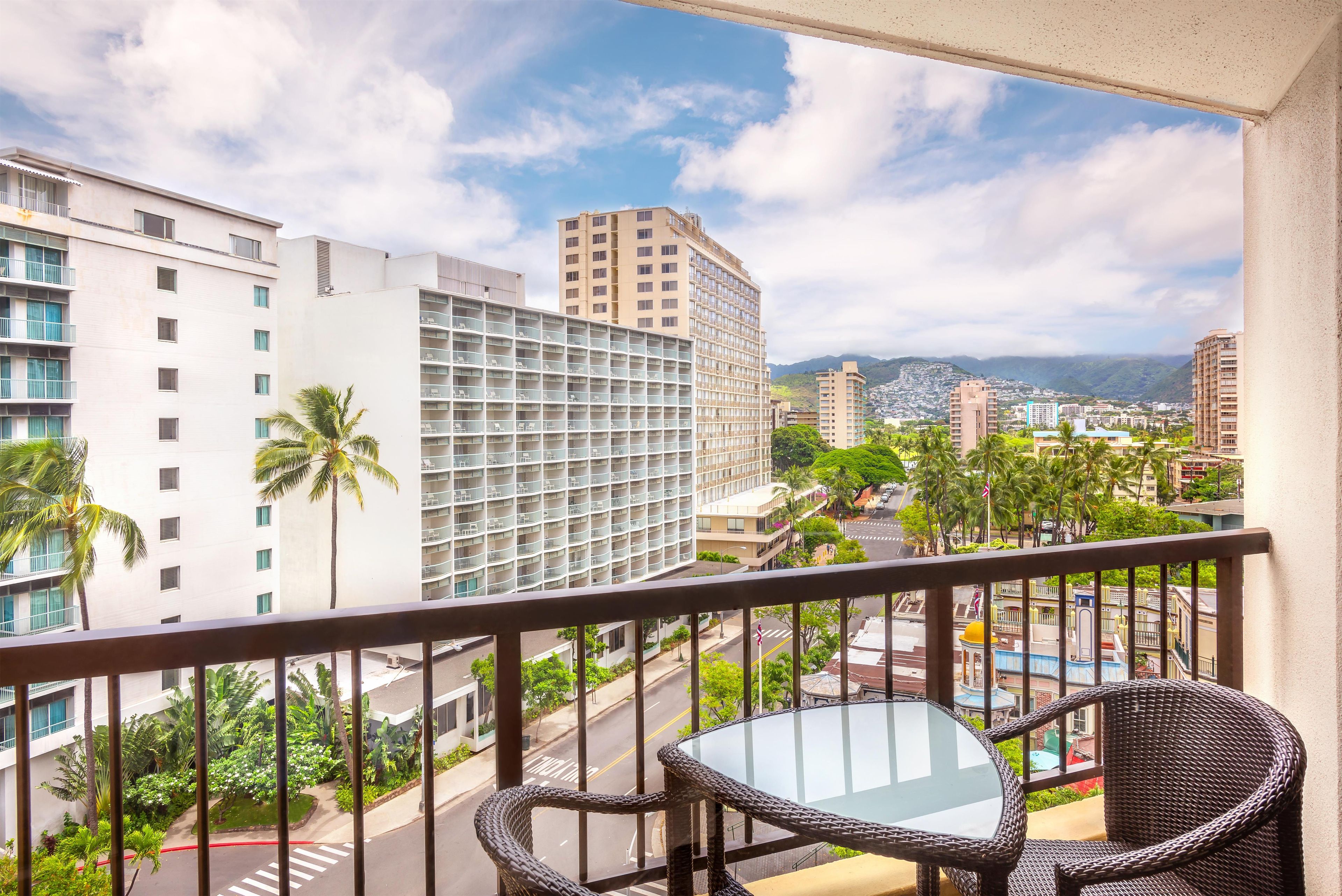 A balcony with wicker chairs and a glass table, overlooking a cityscape with buildings and palm trees.