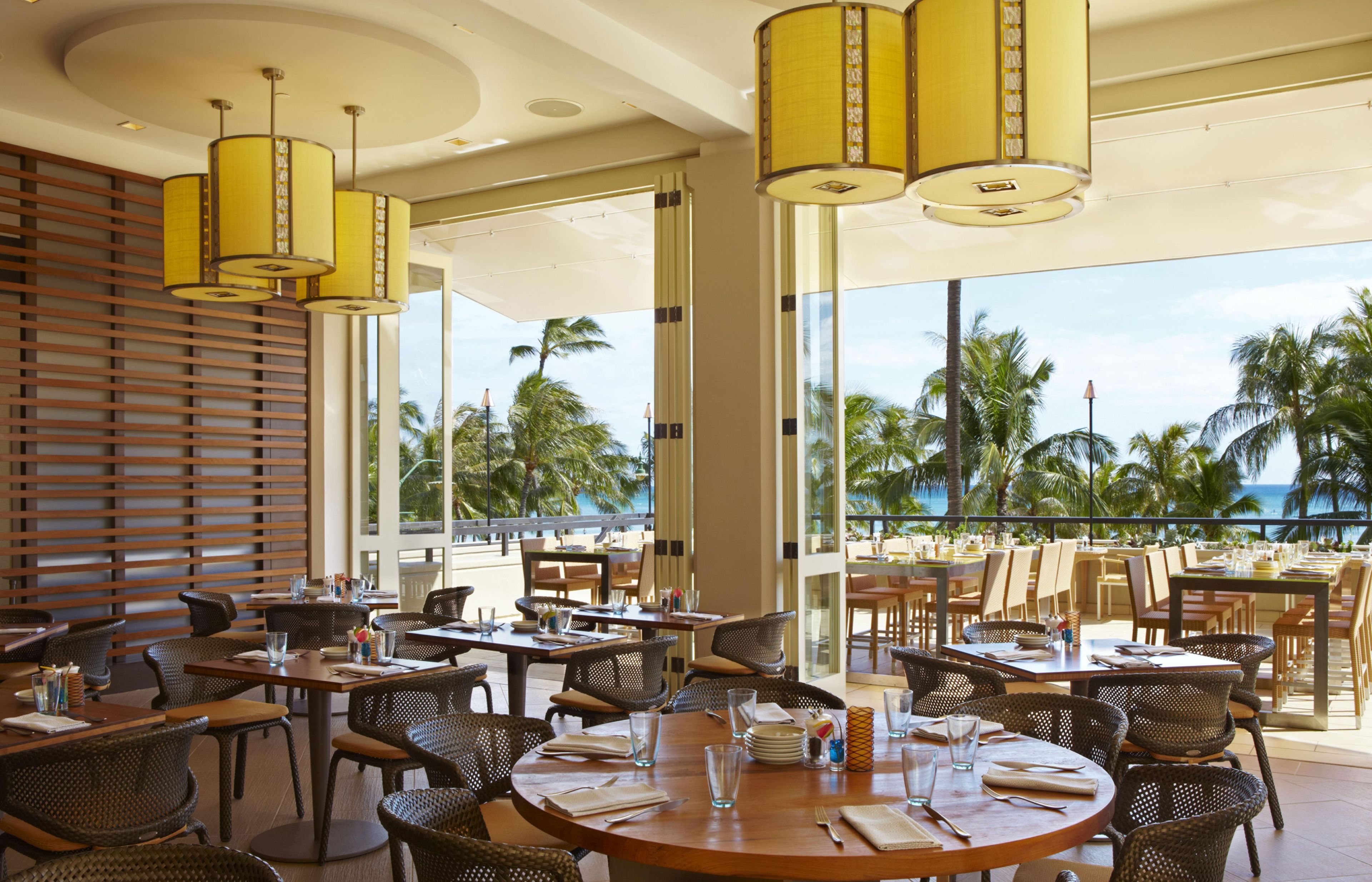 A restaurant with round tables, wicker chairs, and yellow pendant lights.