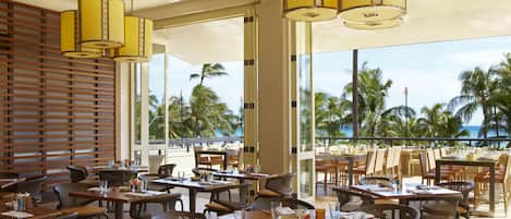 A restaurant with round tables, wicker chairs, and yellow pendant lights.