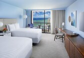 A hotel room with two beds, a desk, a chair, a TV, and a balcony with a view of the ocean and buildings.
