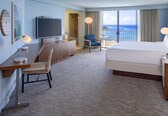 A hotel room with a large bed, a desk with a chair, a TV, and a view of the ocean.