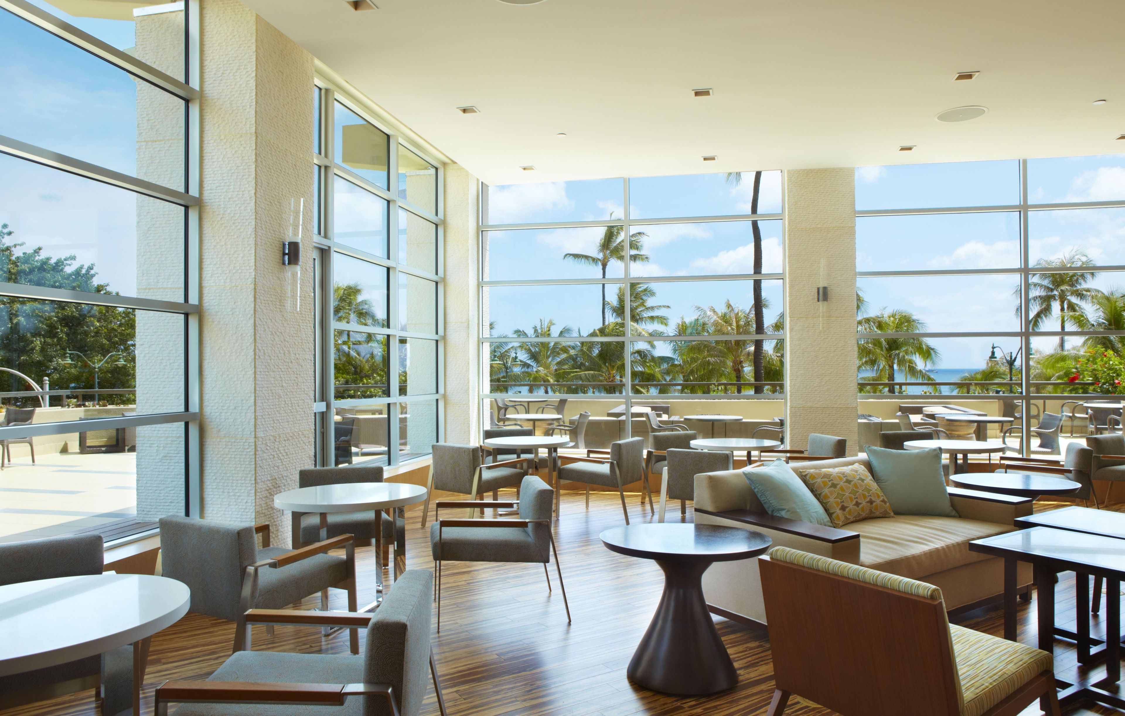 A spacious indoor area with large windows, multiple seating arrangements, and a view of palm trees and the ocean.