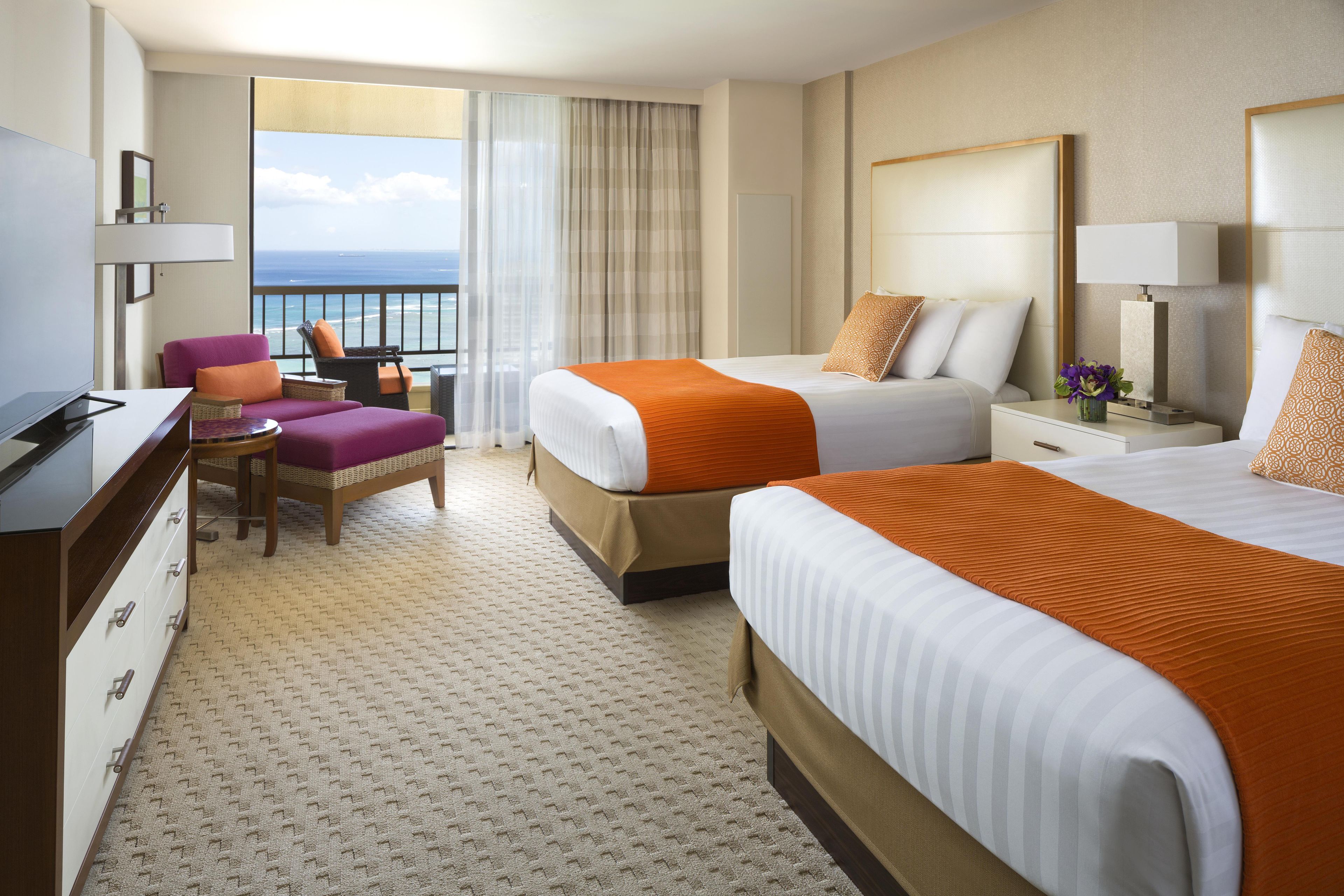 A hotel room with two beds, a desk, a chair, a TV, and a view of the ocean.