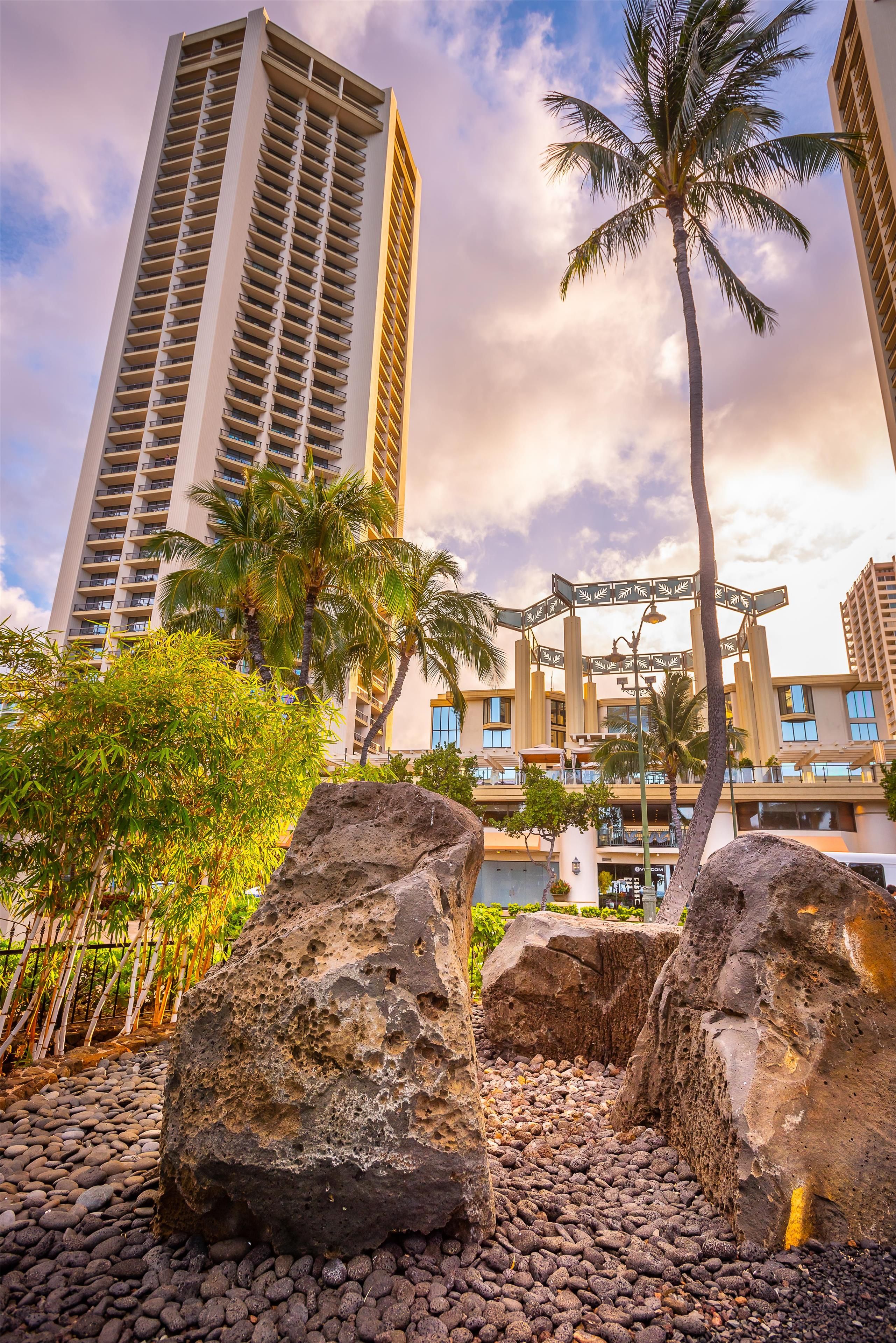 A high-rise building with balconies, palm trees, and a rocky area with large boulders.
