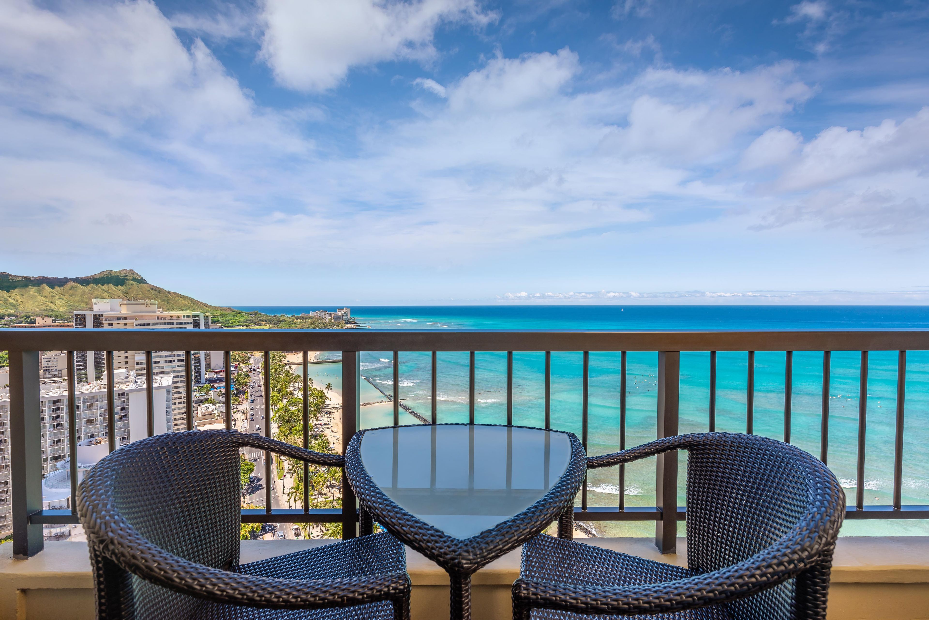 A balcony with two wicker chairs and a glass table overlooking a beach and ocean.
