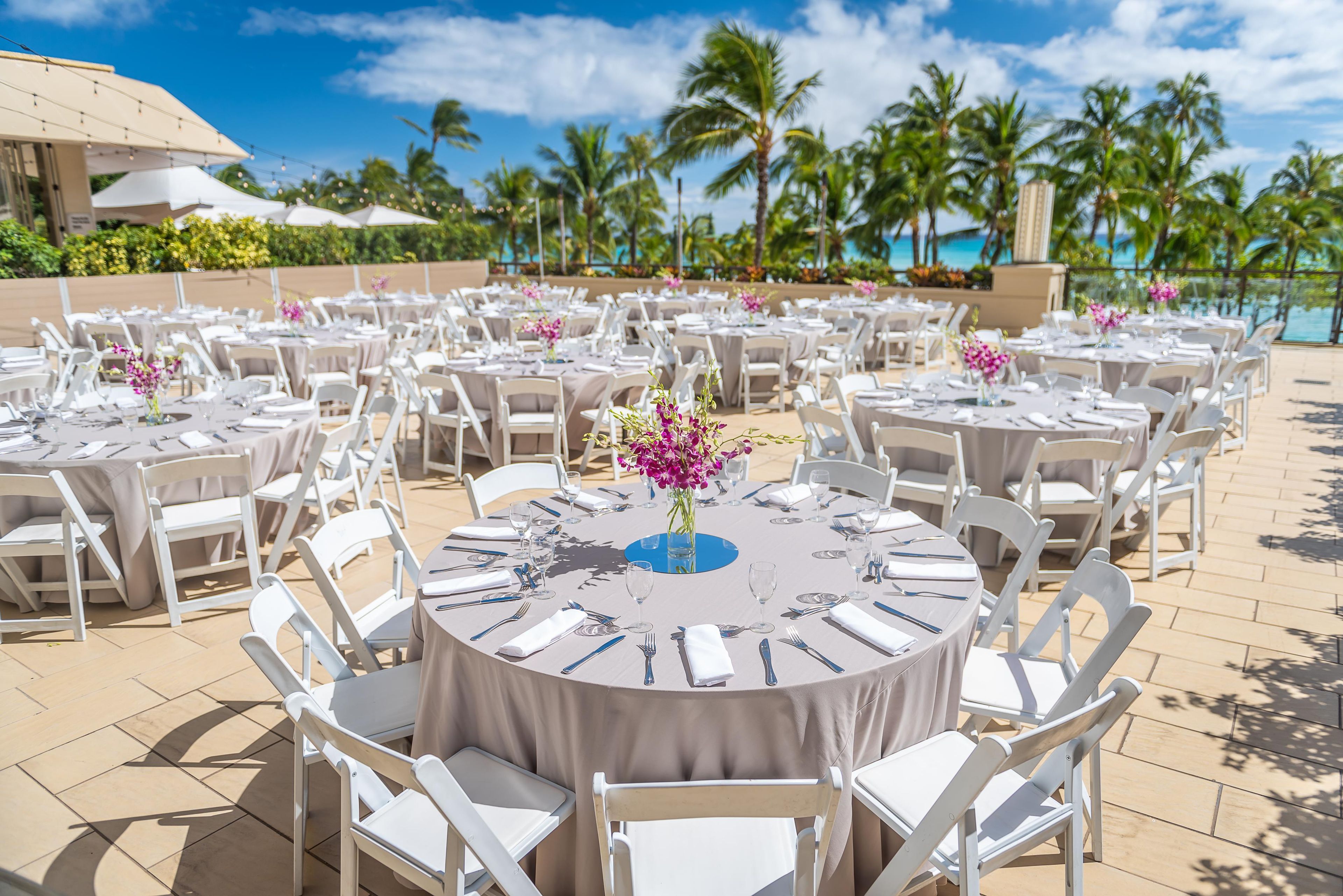 Outdoor round tables with white chairs, arranged in rows, set up for dining.