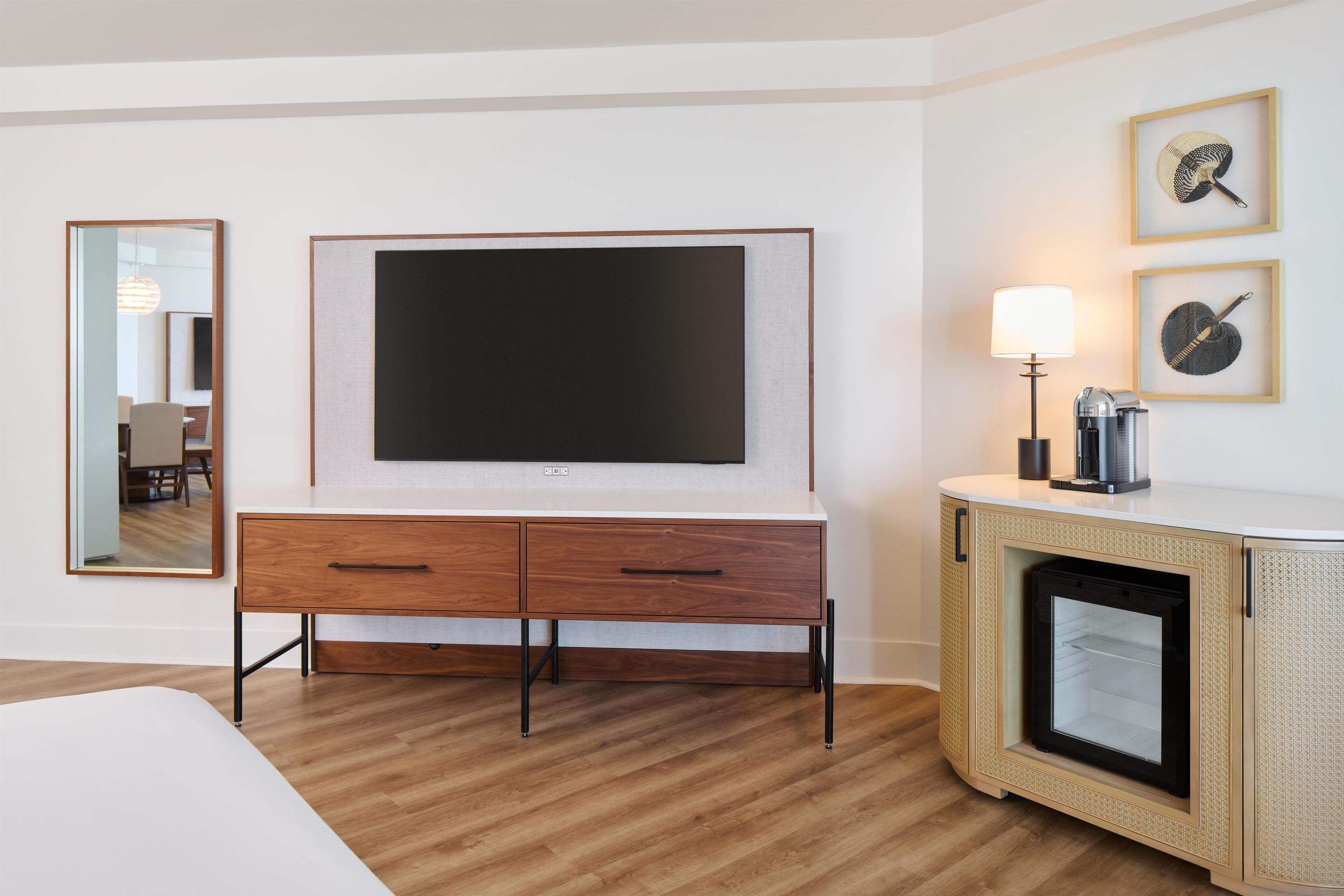 Habitación de hotel con una televisión de pantalla plana, cómoda de madera, refrigerador pequeño, lámpara de noche y cuadros en la pared.