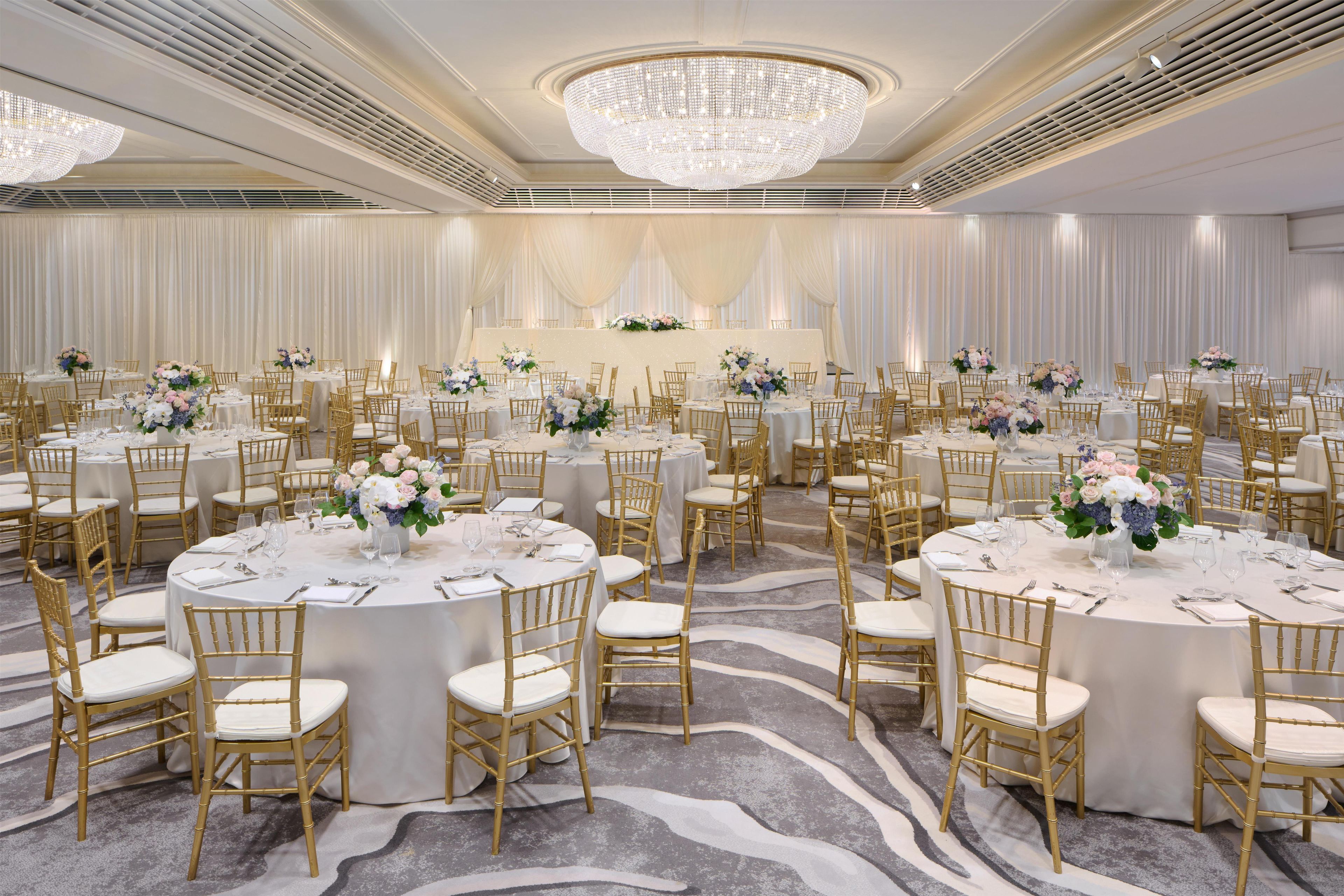 A banquet hall with round tables, gold chairs, and a chandelier.