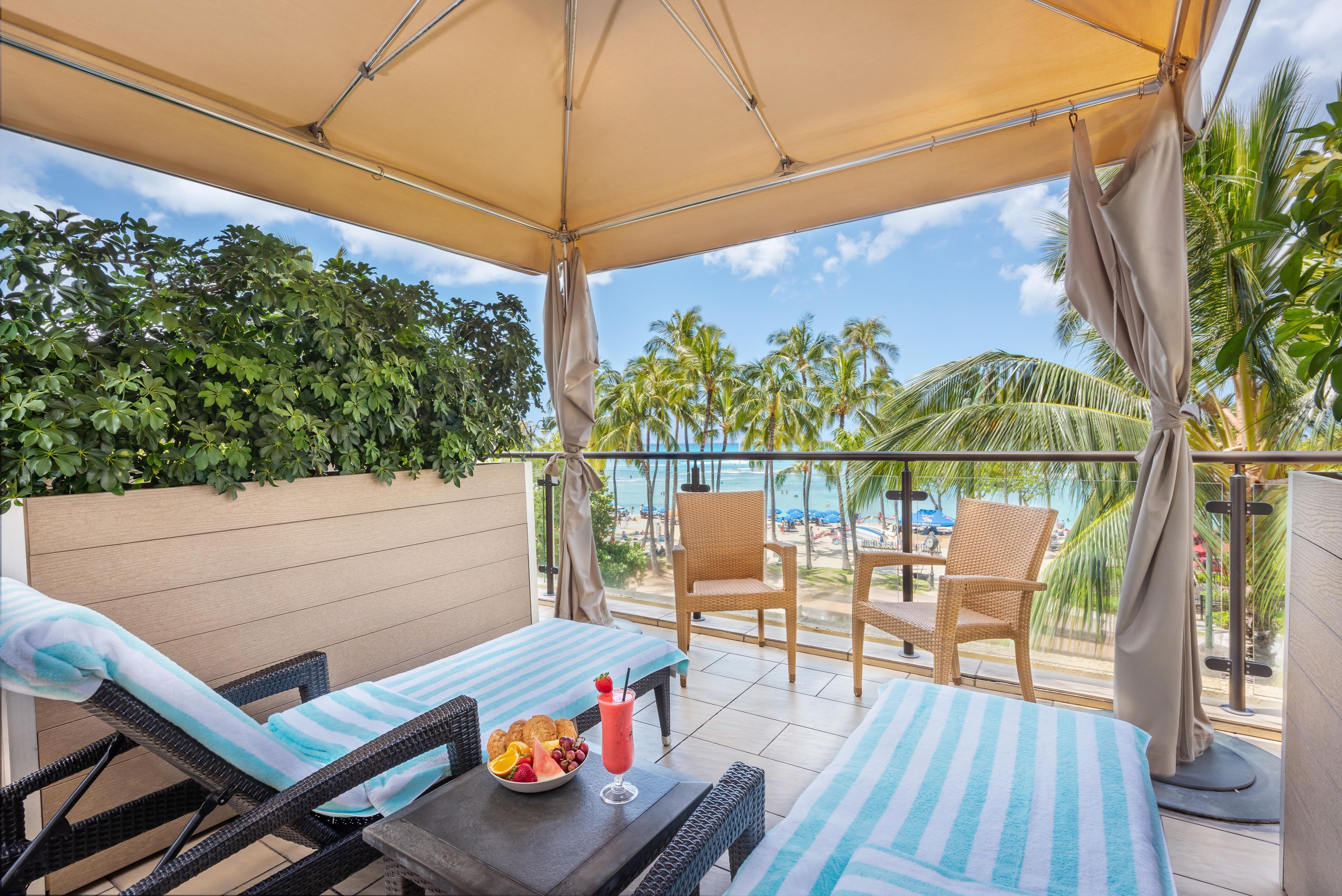 A balcony with wicker furniture, a table with fruit and a drink, and a view of the beach and palm trees.