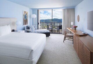 A hotel room with a large bed, a desk, a chair, a balcony with a city view, and a large window.