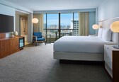 A hotel room with a large bed, a TV, a desk, a chair, a balcony with a view of the city, and a shower.