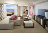A modern hotel room with a large sofa, a coffee table, a TV, and a balcony with a view of the ocean.